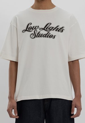 Osoba nosząca luźną białą koszulkę z czarnym kursywnym napisem „Low Lights Studios”, w połączeniu z ciemnymi spodniami.