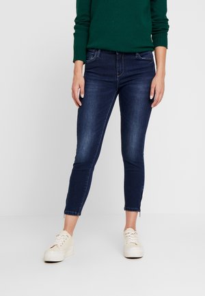 Person trägt dunkelblaue Skinny Jeans mit Reißverschlüssen am Knöchel, einen grünen Langarmpullover und cremefarbene Plateau-Sneaker und steht auf einem weißen Boden.