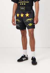 Melnas šortus ar dzeltenu leopardu izdruku un trim sarkaniem zvaigznēm. Materiāli ietver gludu tekstūru, elastīgu vidukļa joslu un adidas logo.
