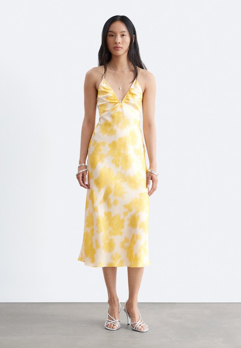 Femme aux longs cheveux foncés porte une robe midi sans manches jaune et blanche fleurie, avec de fines bretelles et des sandales argentées à talons hauts.