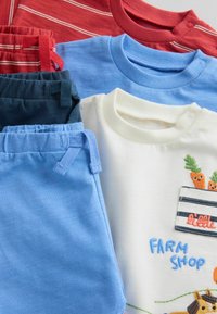 Pile de vêtements pour enfants pliés, comprenant un pantalon bleu, une chemise blanc crème avec une broderie de carotte, ainsi que des hauts rouges et bleu marine à rayures.
