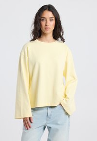 Woman with long dark hair wearing a loose pale yellow long-sleeve shirt and light blue jeans, standing with one hand in pocket.