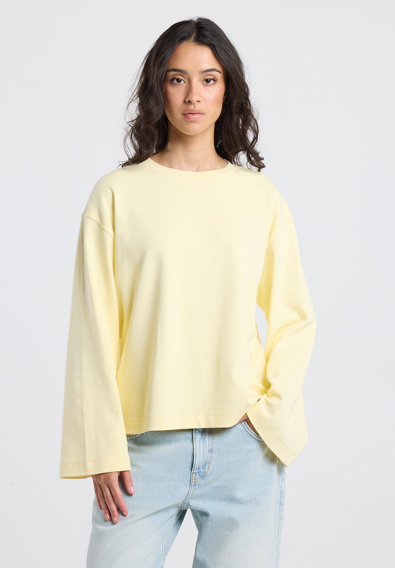 Woman with long dark hair wearing a loose pale yellow long-sleeve shirt and light blue jeans, standing with one hand in pocket.