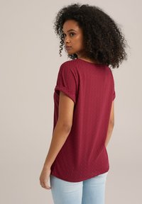 Burgundi värvi tekstuuriga pluus lühikeste rullitud varrukatega, millel on tiku-muster, ümar allääre ja pehme, kerge kangas.