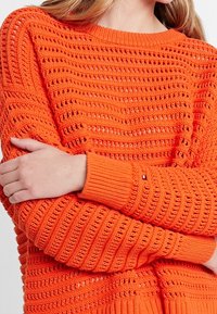 Femme portant un pull tricoté orange vif avec un motif de mailles ouvertes horizontales, croisant les bras sur la poitrine.