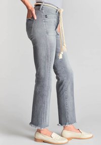 Jeans en denim gris clair avec une coupe droite, ourlet effiloché, cinq poches et une ceinture en corde. Associés à des mocassins crème, montrant un style décontracté.