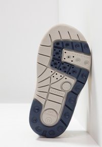 Podešev boty s texturovaným povrchem; obsahuje kombinaci béžové a námořnické barvy, ventilační otvory, vycpávané části a detaily značky.