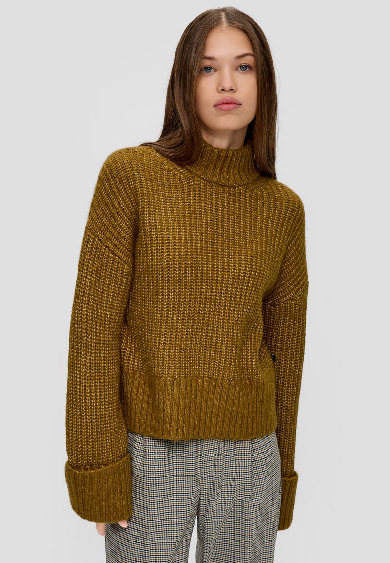 Senfgelbes Strickpullover mit hohem Kragen, lockerer Passform und breiten Bündchen; strukturierter Stoff mit geripptem Saum. Kombiniert mit karierter Hose.