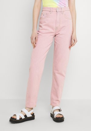 Persoon die lichtroze jeans met hoge taille, een tie-dye top en witte platformsandalen met zwarte zolen draagt, staand tegen een witte achtergrond.