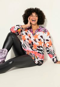 Bunte Langarmbluse mit einem Mischmuster aus Sternen, Streifen und Blumen in Pink, Orange und Schwarz, kombiniert mit glänzenden schwarzen Leggings.