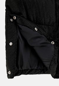 Chaqueta negra acolchada, con patrones de puntos brillantes, cierres a presión y un forro interior suave. La textura es blanda, con un diseño ligeramente acolchado.
