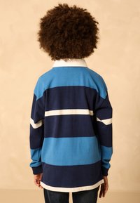 Mėlynas ir tamsiai mėlynas dryžuotas ilgomis rankovėmis marškinėliai su balta apykakle, pasižymintys laisvu kirpimu ir minkšta tekstūra, dėvimi ant tamsių džinsų.