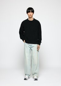 Ung mann iført en svart strikket genser, lyseblå vidde jeans og hvite joggesko, som står mot en ensfarget hvit bakgrunn.
