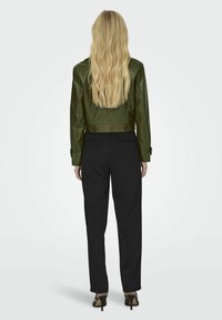 Giacca di pelle verde oliva con dettagli a bottoni, abbinata a pantaloni neri a vita alta. Capelli lunghi biondi visibili.