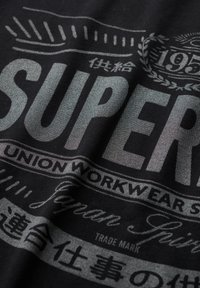 Zwarte stof met zilveren tekst met onder andere "SUPER", "UNION WORKWEAR", "195" en Japanse karakters, ontworpen in een vintage, verweerde stijl.