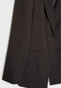 Blazer marron foncé cintré avec une fermeture à un bouton et deux poches avant horizontales sur un fond clair.