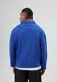 Kobaltblauwe pulloverjas met een brede kraag en lange mouwen, gemaakt van zachte stof, gezien vanaf de achterkant, gecombineerd met een zwarte broek.