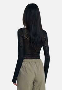 Top nero a maniche lunghe con motivo a onde, realizzato in materiale trasparente. Abbinato a pantaloni leggeri color khaki. Vista posteriore che mostra il design aderente.