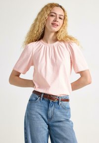 Helles rosa Kurzarmoberteil mit gerafftem Ausschnitt und Spitzenbesatz, kombiniert mit hochtaillierten blauen Jeans und einem braunen Ledergürtel.