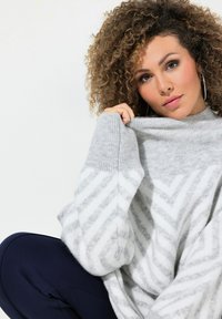 Pull en laine gris avec des rayures blanches en chevron, coupe ample, poignets côtelés et col montant. Modèle assis, tenant le col du pull.