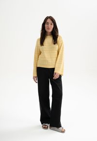 Geel gestructureerd sweater met een ronde hals, gecombineerd met zwarte wijde broek en grijze sandalen, staand op een witte achtergrond.