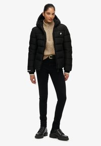 Sort pufferjakke med hætte, har en frontlynlås og hvidt logo på brystet. Bæres over en beige striktrøje og sorte skinny jeans.