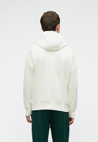 Homem a usar um hoodie branco simples e calças verde-escuro, visto de costas contra um fundo branco liso.