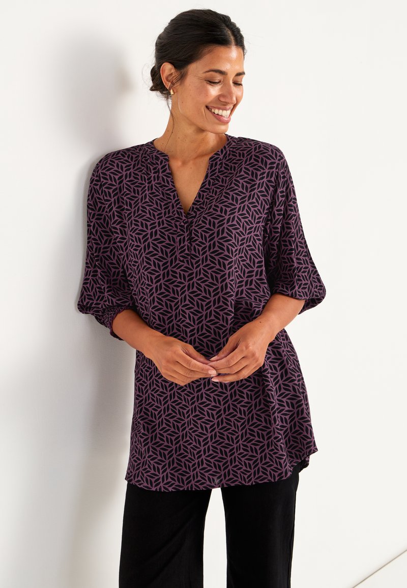 Paarse tuniek met geometrisch bladenpatroon, V-hals en 3/4 mouwen. De stof lijkt zacht met een lichte textuur, gecombineerd met zwarte broek.