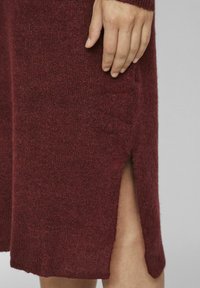 Main reposée sur une robe en tricot bordeaux arrivant au genou, avec une fente sur le côté, montrant une partie de la jambe inférieure sur un fond gris.
