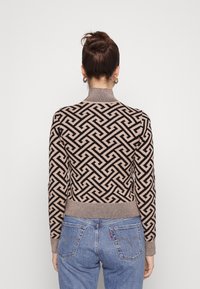 Pull-over tricoté beige avec un motif géométrique noir. Il présente un col montant et des poignets côtelés, associé à un jean en denim bleu.