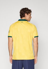 Mann som står med ryggen til, iført en gul kortermet skjorte med grønn krage og mansjetter, samt marineblå shorts mot en enkel grå bakgrunn.