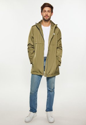 Olijfgroene waterdichte jas met ritssluiting, verstelbare capuchon en voorzakken, gecombineerd met lichtblauwe spijkerbroek en witte sneakers.