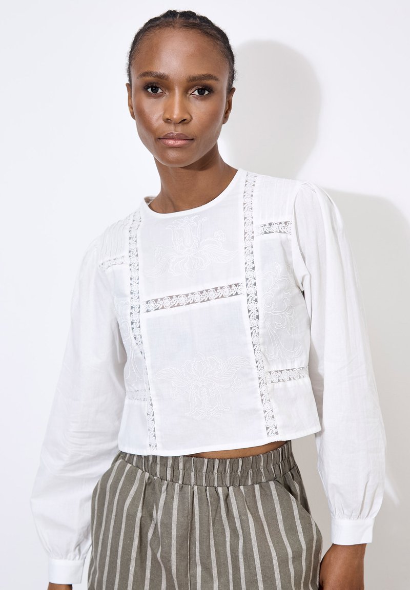Witte cropped blouse met lange mouwen, ingewikkelde borduursels aan de voorkant en kantaccenten. Gecombineerd met gestreepte, elastische taille broek.