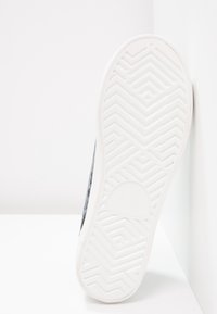 Semelle d'une basket blanche avec un motif de bande de roulement géométrique, posée contre un mur d'angle blanc sur une surface blanche.