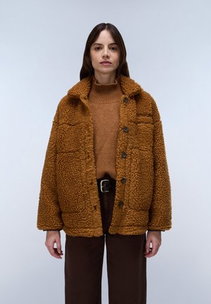 Kvinde iført brun, vamset jakke med knapper, brun rullekravetrøje og mørke bukser med sort bælte, stående mod en enkel lys baggrund.
