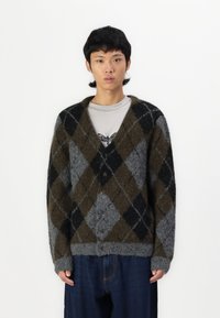Cardigan argyle grigio, marrone e nero con una texture morbida, chiusura a bottoni e taglio oversize, abbinato a una camicia grigia e pantaloni di denim.