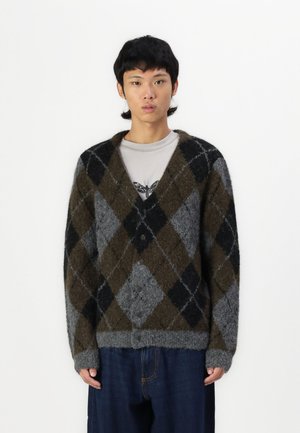 Cardigan argyle grigio, marrone e nero con una texture morbida, chiusura a bottoni e taglio oversize, abbinato a una camicia grigia e pantaloni di denim.