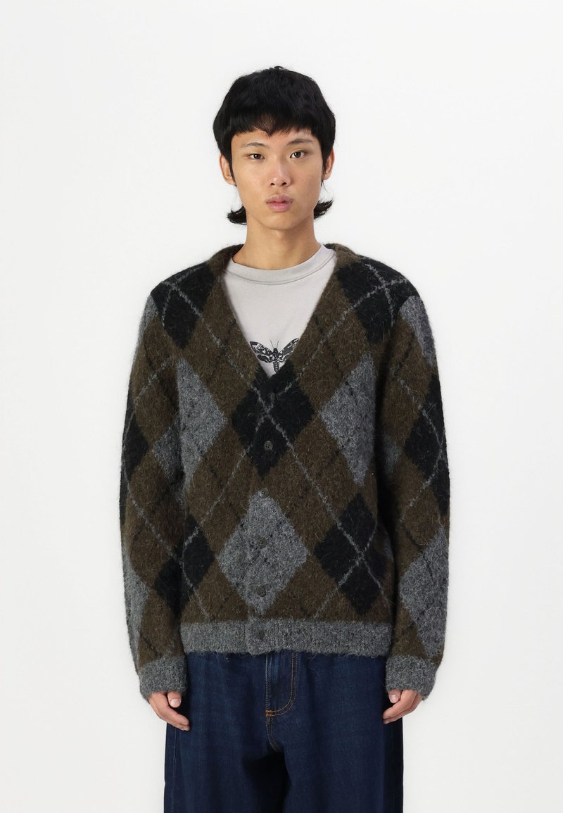 Cardigan argyle grigio, marrone e nero con una texture morbida, chiusura a bottoni e taglio oversize, abbinato a una camicia grigia e pantaloni di denim.
