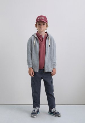 Grijze rits hoodie met koordjes, bordeaux polo shirt daaronder, donkere cargobroek en grijze sneakers met blauwe accenten. Rode pet op het hoofd.