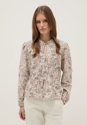 Jonge vrouw die een crèmekleurige blouse met paisley-patroon en lange mouwen draagt, met crèmekleurige broek tegen een effen achtergrond.