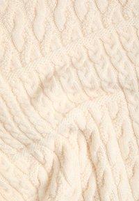 Tissu tricoté de couleur crème avec des motifs tressés et torsadés complexes, montrant une texture douce et des sections pliées.