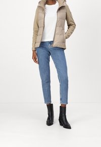 Béžová péřová bunda s kapucí, střih do pasu, zapínání na zip, v kombinaci se světle modrými rovnými džíny a černými botami na podpatku kotníkového střihu.