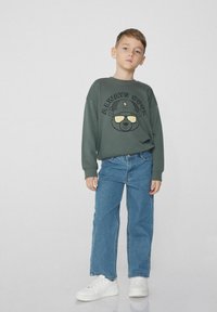 Tmavě zelená mikina s grafikou medvěda "Always Cool", v kombinaci se světle modrými džíny a bílými teniskami. Střih je volný a neformální.