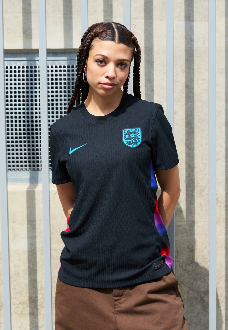 Tricou negru Nike cu mâneci scurte, prezintă o textură orizontală, logo-ul teal și emblema Angliei. Panourile laterale afișează un gradient de la roșu la violet.