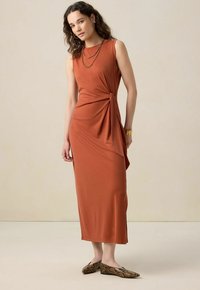 Robe sans manches couleur rouille avec un détail torsadé sur le côté, en tissu doux, associée à des ballerines imprimé serpent sur un fond neutre.