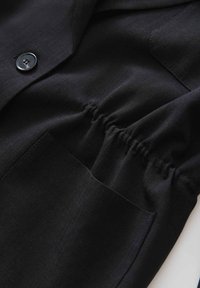 Zwarte stof met een gladde textuur, voorzien van een knoopsluiting en elastische gathers op de taille. Inclusief een zijzakdetail.