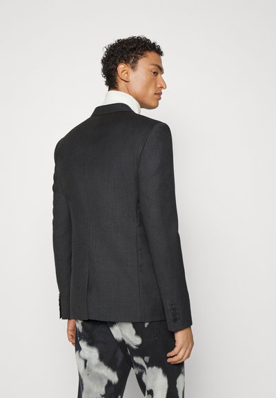 Dunkelgrauer Blazer mit strukturiertem Design, Reverskragen und weißem Kragen. Texturiertes Material und Details mit drei Knöpfen an den Ärmeln, getragen über gemusterten Hosen.