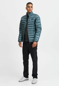 Teal pufferjack met een rits aan de voorkant, hoge kraag en horizontale stiksels. Zwarte jeans en lichtgrijze sneakers maken de look compleet.