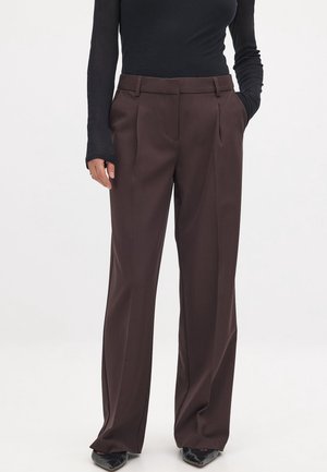 Kvinde iført mørkebrune, viddebukser med højtaljet talje og forreste folder, kombineret med en tætsiddende sort langærmet top og sorte spidse sko.