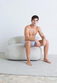 Gekrümmtes, weiches, hellgraues Sofa mit samtiger Textur. Eine sitzende Person in blauen Unterhosen auf einem cremefarbenen Teppich, vor einem schlichten weißen Hintergrund.